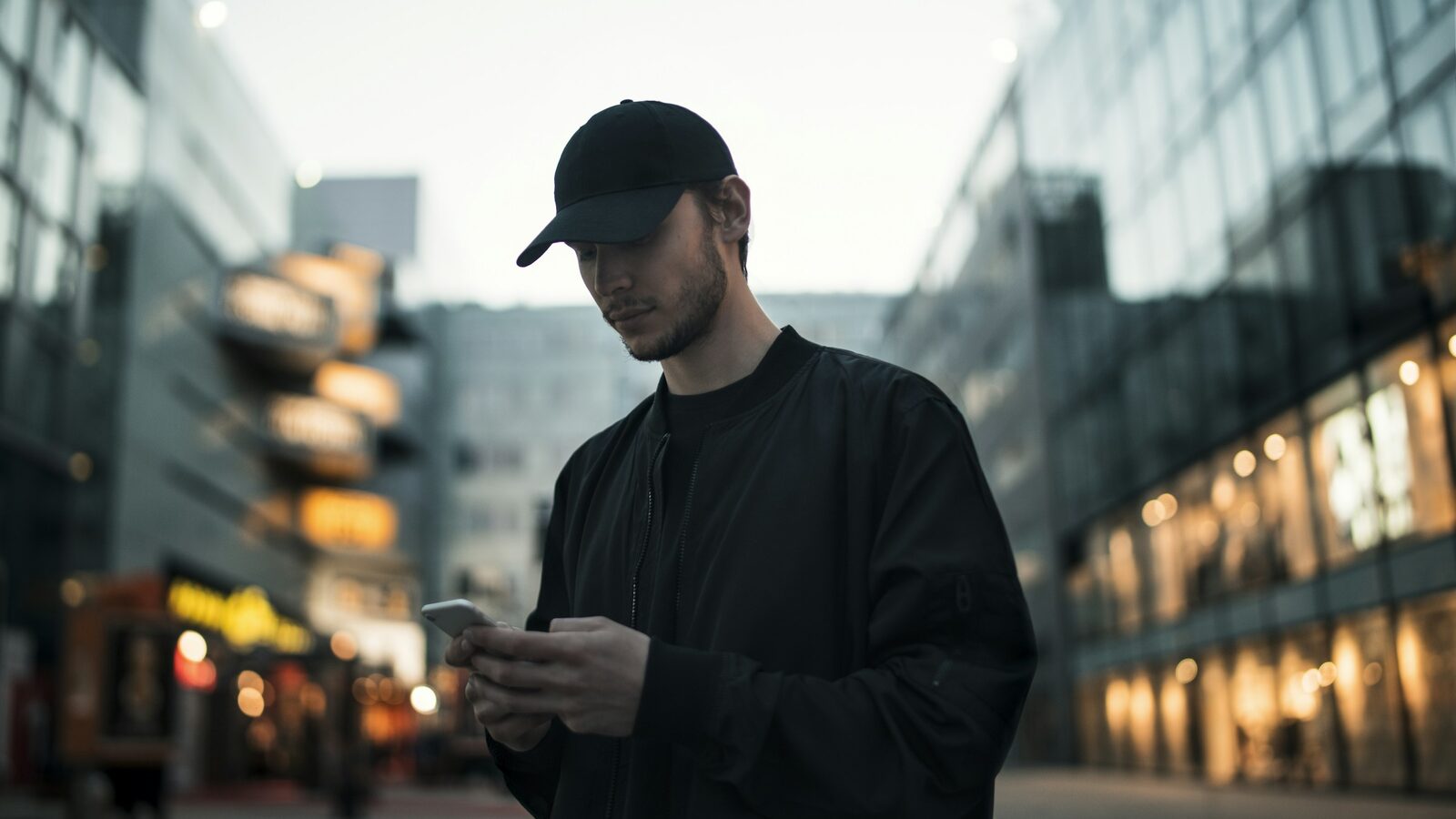 Mann i svart jakke og svart caps står i en bygate i skumringen og ser på mobilen sin.
